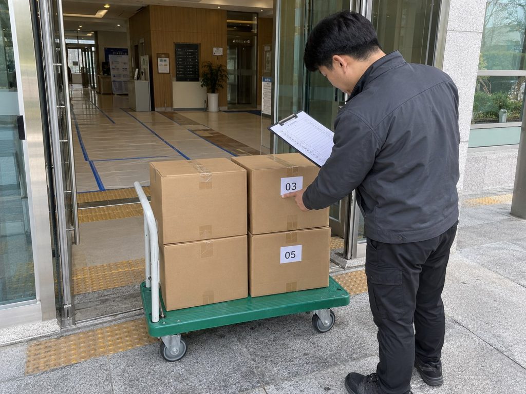 건물 입구에서 점검표를 보며 번호 라벨이 붙은 박스를 확인하는 모습